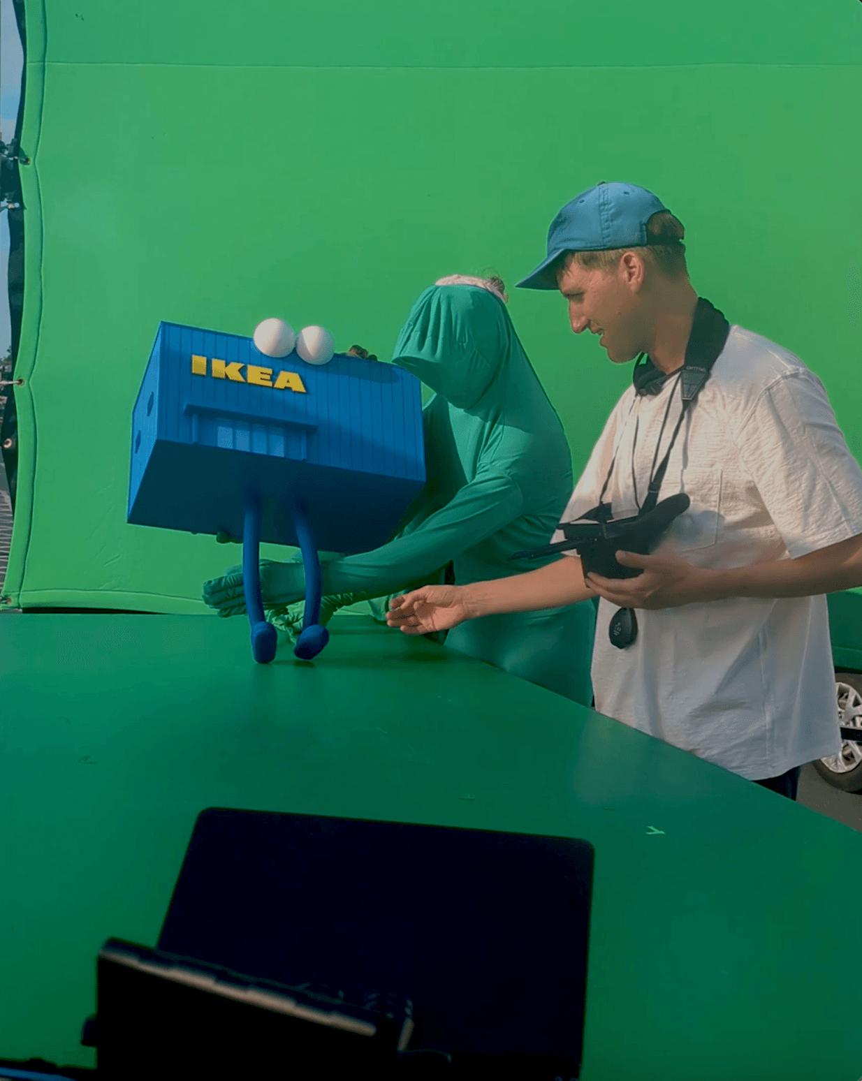 Behind-the-scenes photo from an IKEA commercial shoot, with a puppeteer and director operating a cartoon IKEA building character against a green screen.